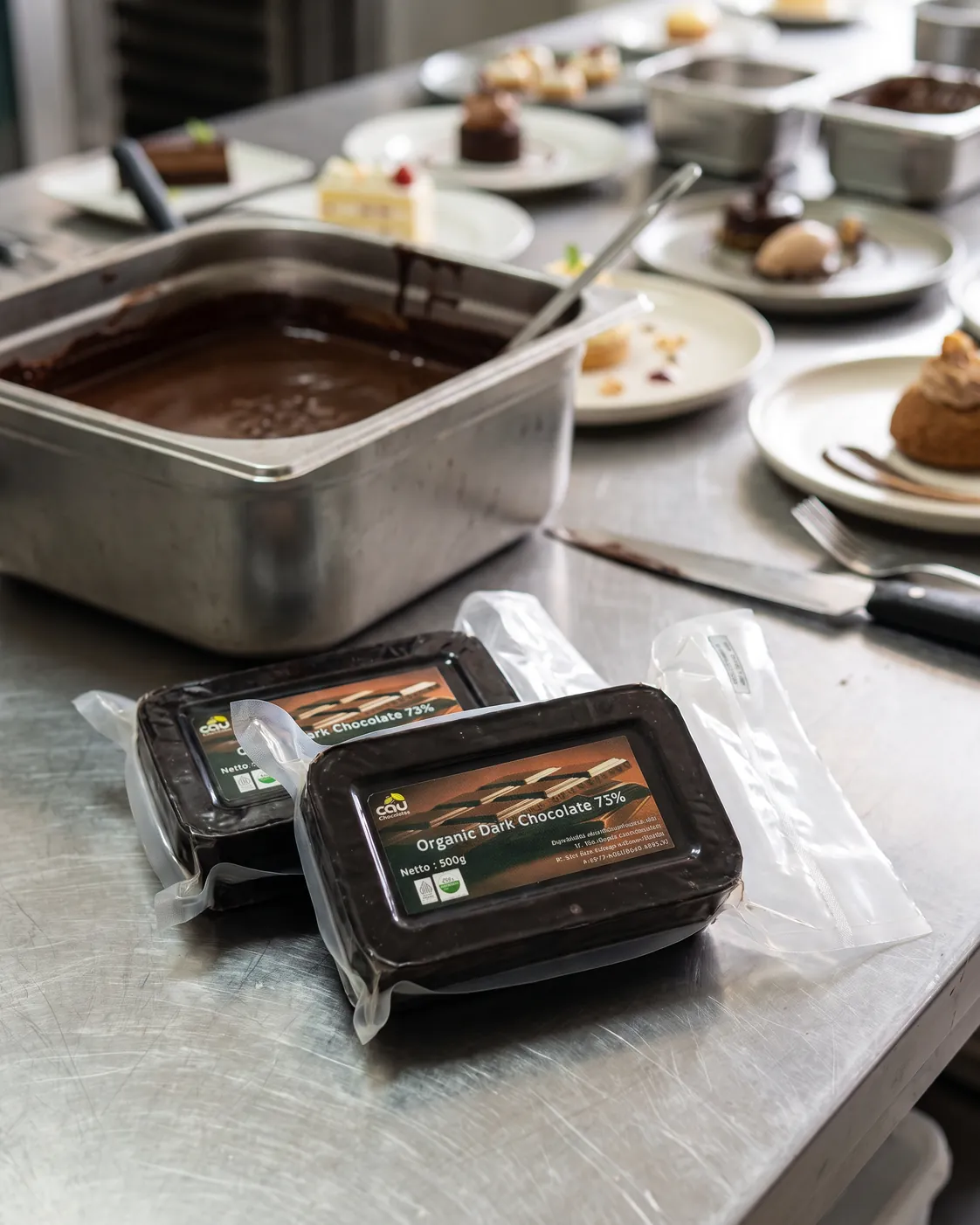 AI visual — CAU Organic Dark Chocolate 73% couverture 500 g packs in a hotel pastry kitchen (real packaging, AI-styled background)